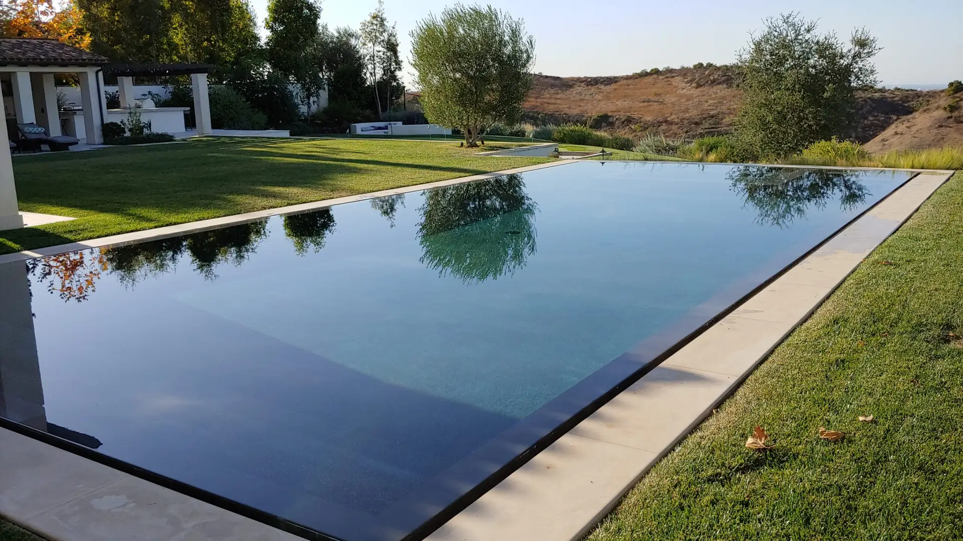 Infinity pool reflecting surrounding trees and clear sky, with a manicured lawn and hillside in the distance.