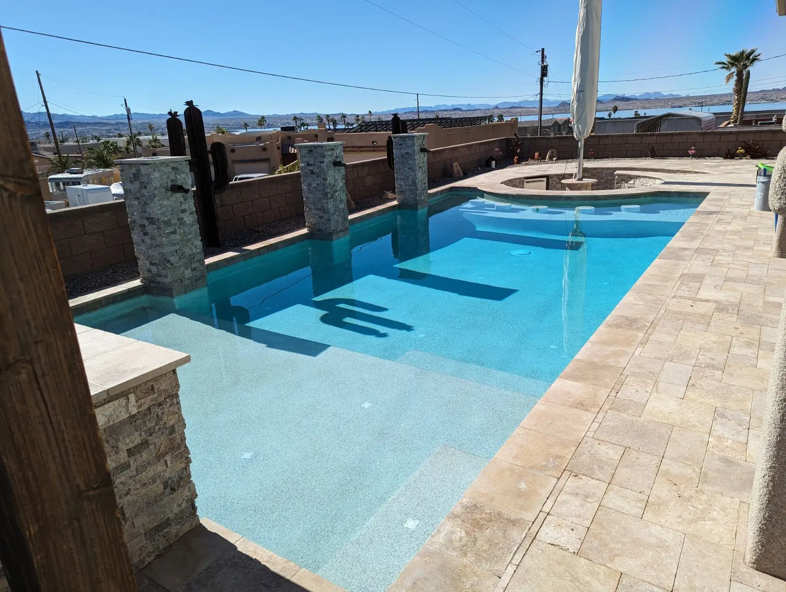 Backyard swimming pool with clear blue water, stone pillars, and a circular spa in a sunny desert setting with mountains in the distance, tiled deck around.