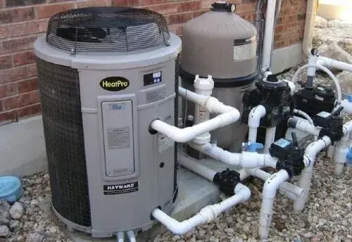 Outdoor HVAC condenser unit with white PVC piping, pool pump, and filtration equipment installed beside a brick wall.