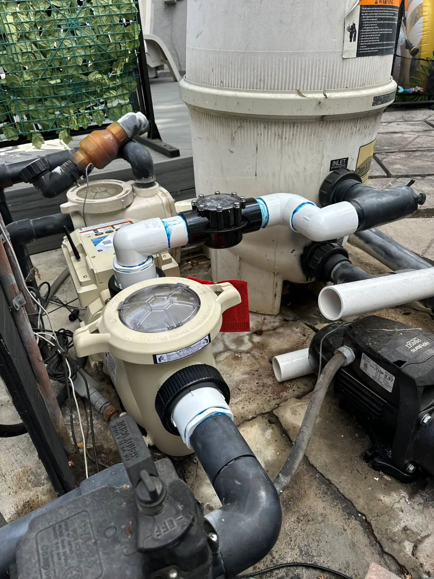 Outdoor pool filtration setup with a white cylindrical filter, beige pump, and black PVC pipes connected.